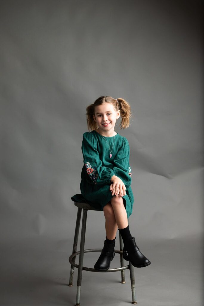 A young girl with a smile, sitting on a stool in a green dress with ankle boots against a gray background.
