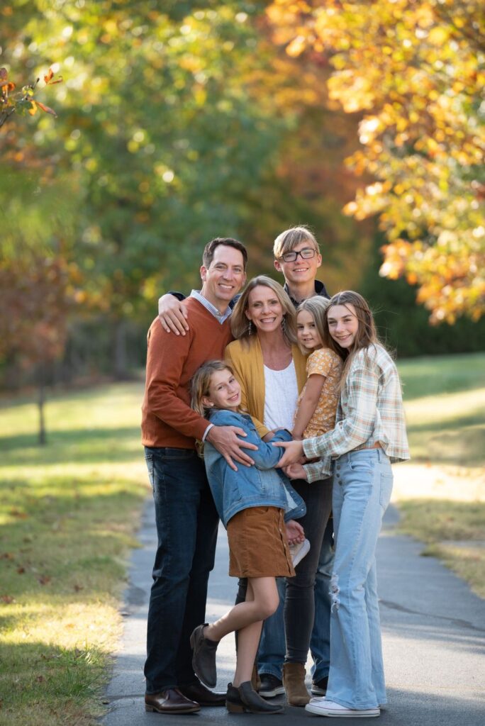 A family of five posing together on a tree-lined path in autumn.