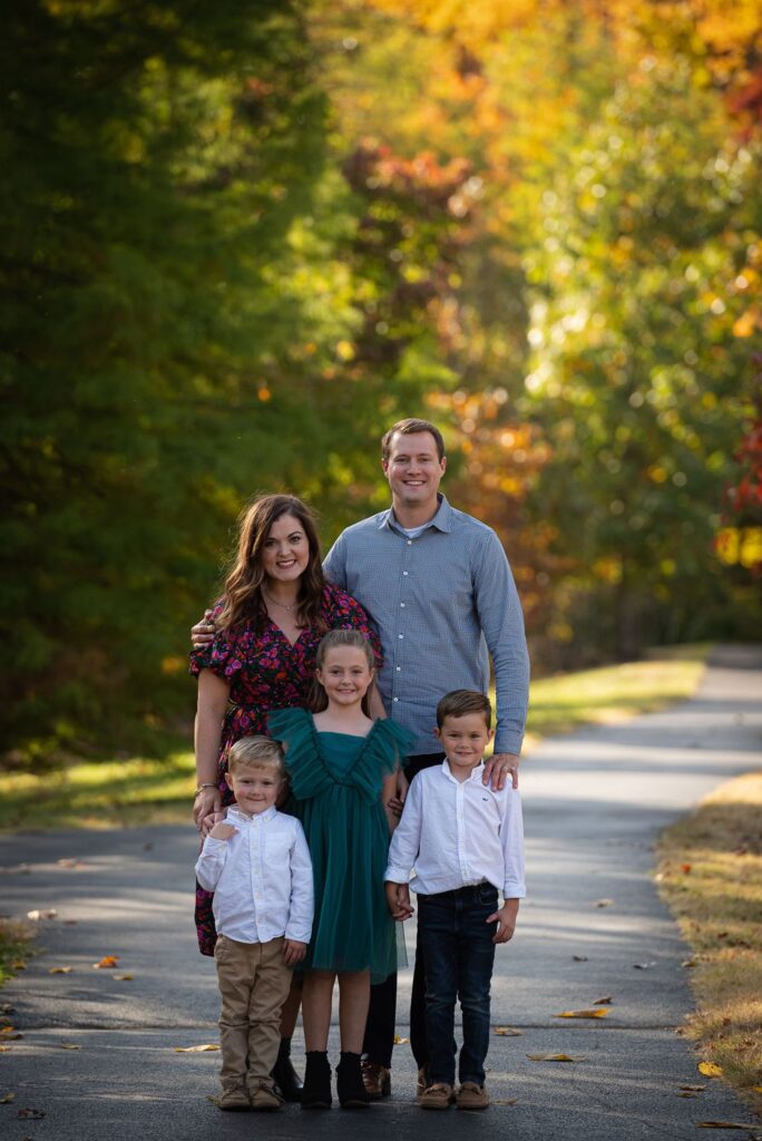 A family of five posing for a photo on a tree-lined path during autumn.