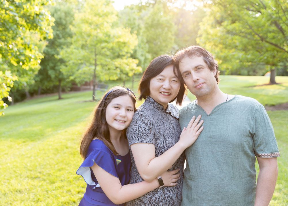 A family of three smiling and embracing each other in a sunlit park.