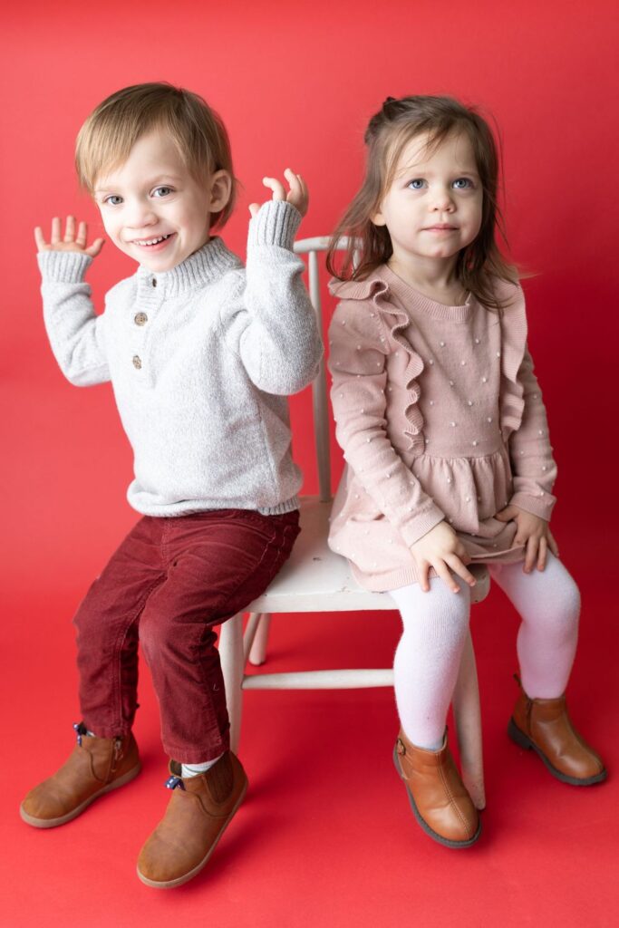 Two young children posing on a white chair against a red background; the boy is waving while the girl sits beside him.