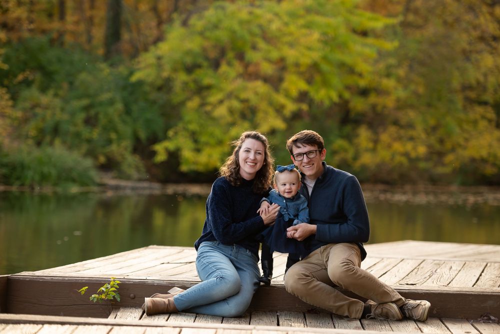 A family with a baby sitting together on a wooden dock against an autumnal backdrop.