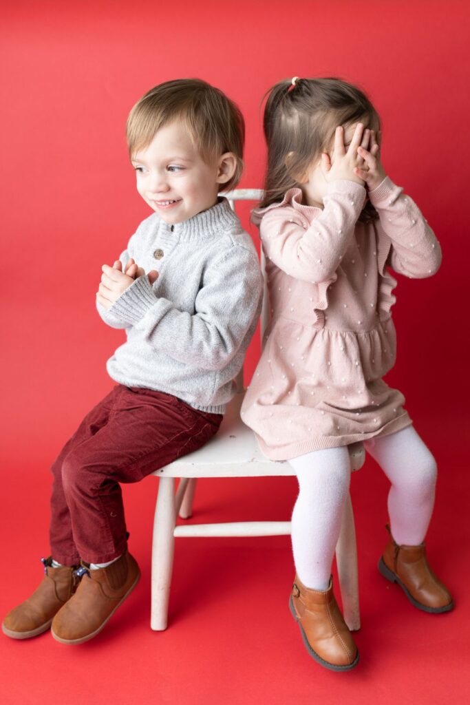 Two young children on a chair; a boy is smiling with crossed arms, and a girl is covering her face with her hands.