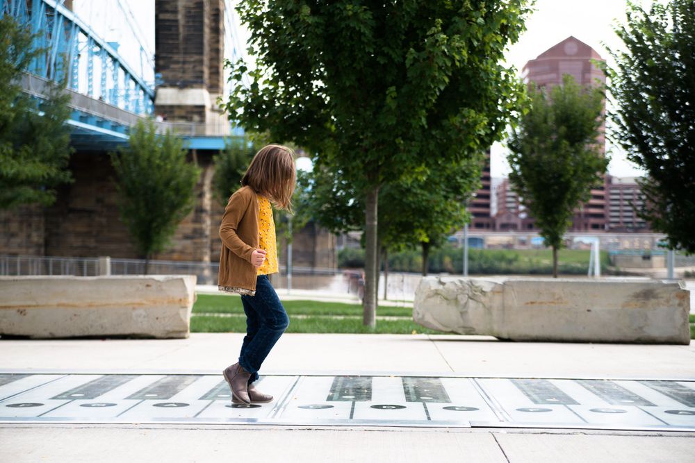 A child in a brown jacket and blue jeans plays on a hopscotch grid in an urban park with trees and a bridge in the background.
