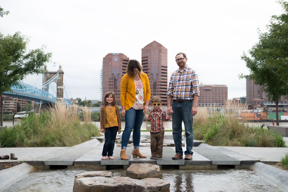 A family of four standing on stepping stones in an urban park with a bridge and buildings in the background.