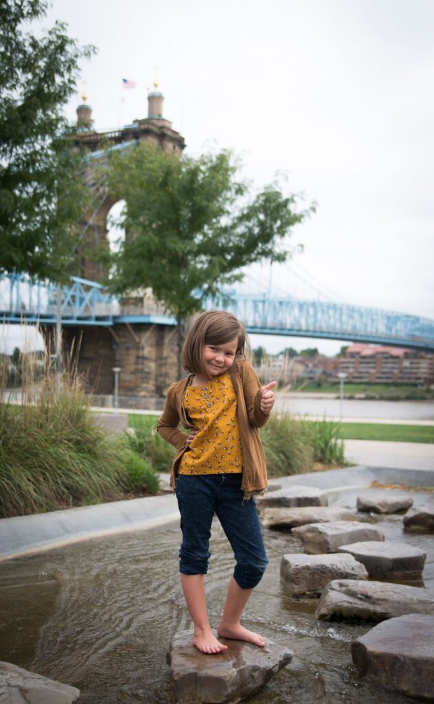 A child balancing on rocks near a river with a bridge in the background.
