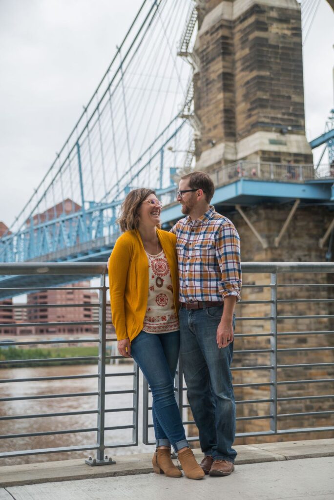 A smiling couple in casual attire standing in front of a suspension bridge.