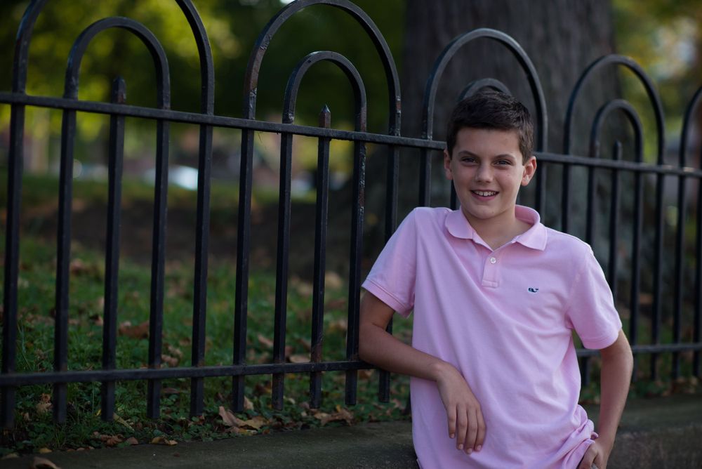 A young boy in a pink polo shirt smiling near a black iron fence.