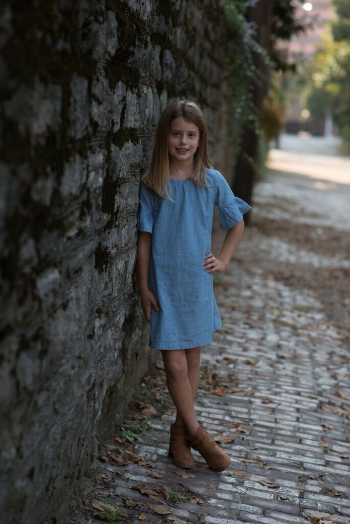 Young girl in a blue dress standing beside a stone wall on a cobblestone path.