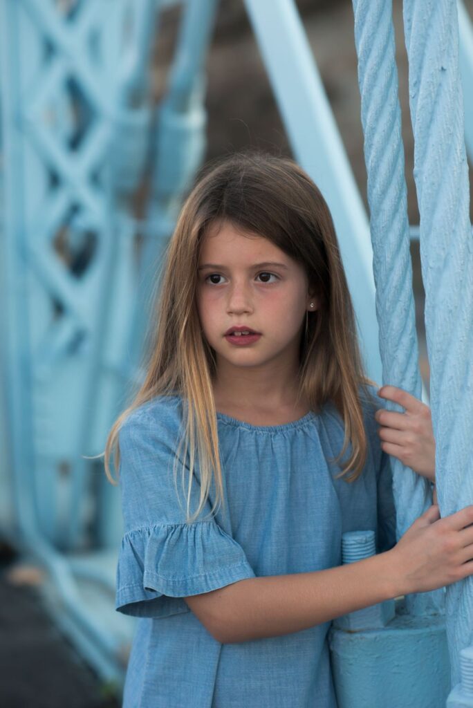 Young girl in a blue dress leaning on a blue bridge railing.