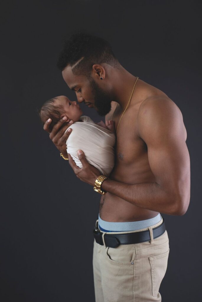 A shirtless man tenderly holding and gazing at a newborn baby against a dark background.