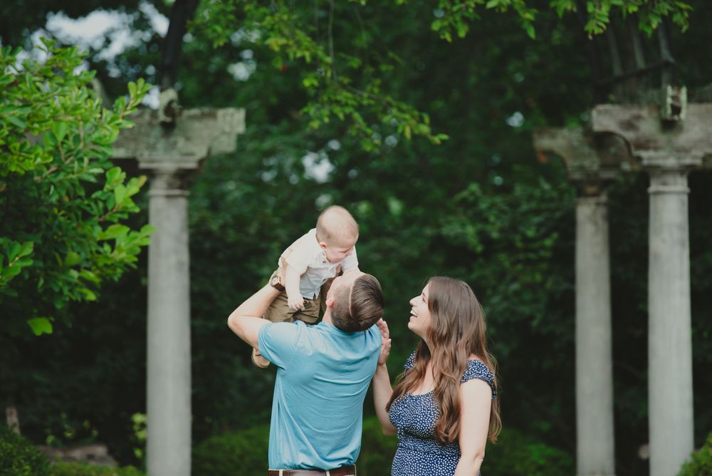 A family enjoys time together outdoors as a parent lifts a baby into the air while the other parent watches with a smile.