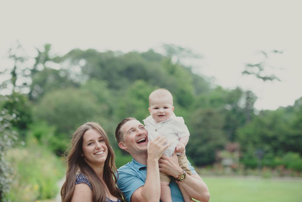 A joyful family moment with a man lifting a baby into the air as a woman smiles beside them, outdoors in a green setting.