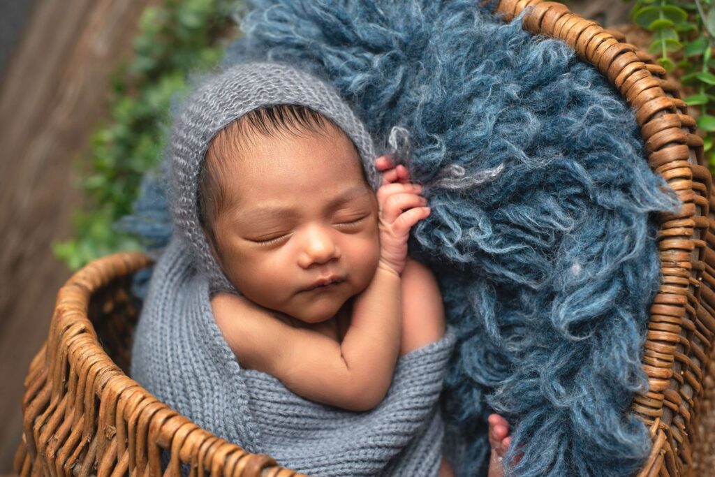 Newborn baby sleeping peacefully in a wicker basket with a blue fuzzy blanket and wearing a knitted gray hat, perfectly arranged by an expert event planner.