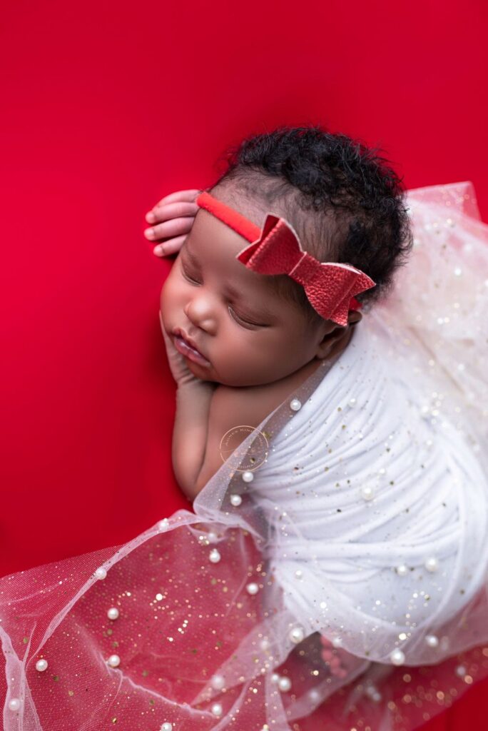 A sleeping infant with a red headband lies wrapped in a white, starry fabric against a red background.