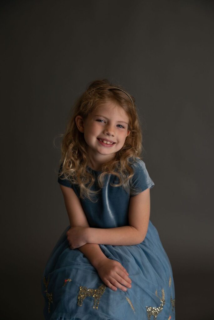 A smiling young girl with curly hair wearing a blue dress poses for a portrait against a dark background.