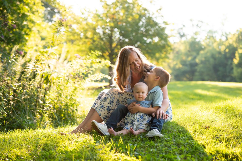 A woman sitting on the grass with two children, smiling in a sunlit park.