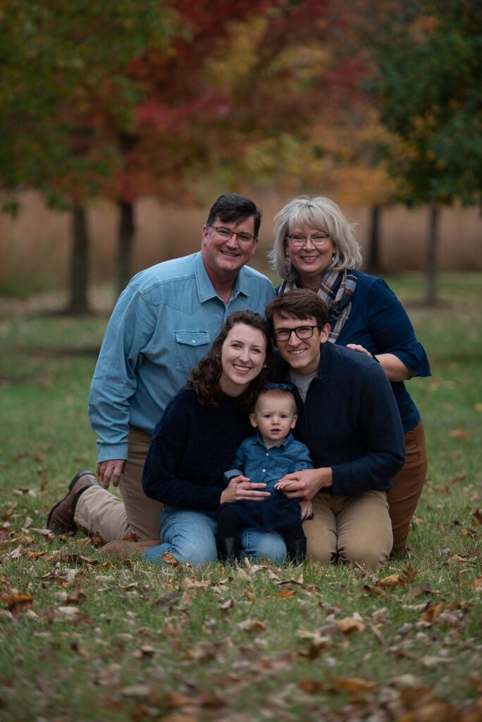 A family of five is posing for a portrait outdoors in a park, with autumn leaves scattered around them.