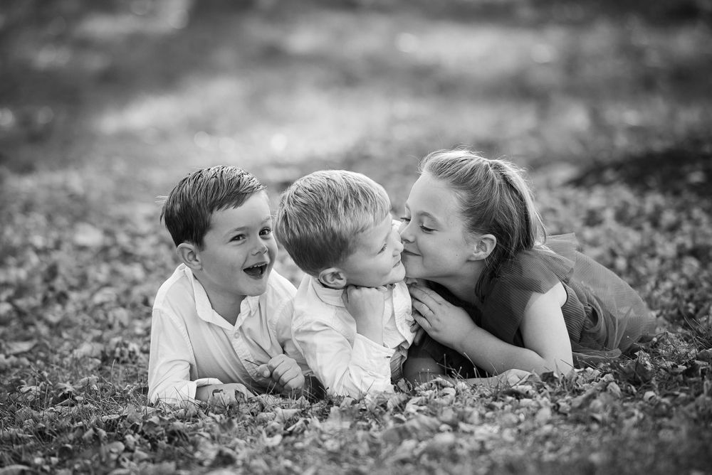 Two children affectionately kissing a smiling young boy on the cheeks while lying on grass.