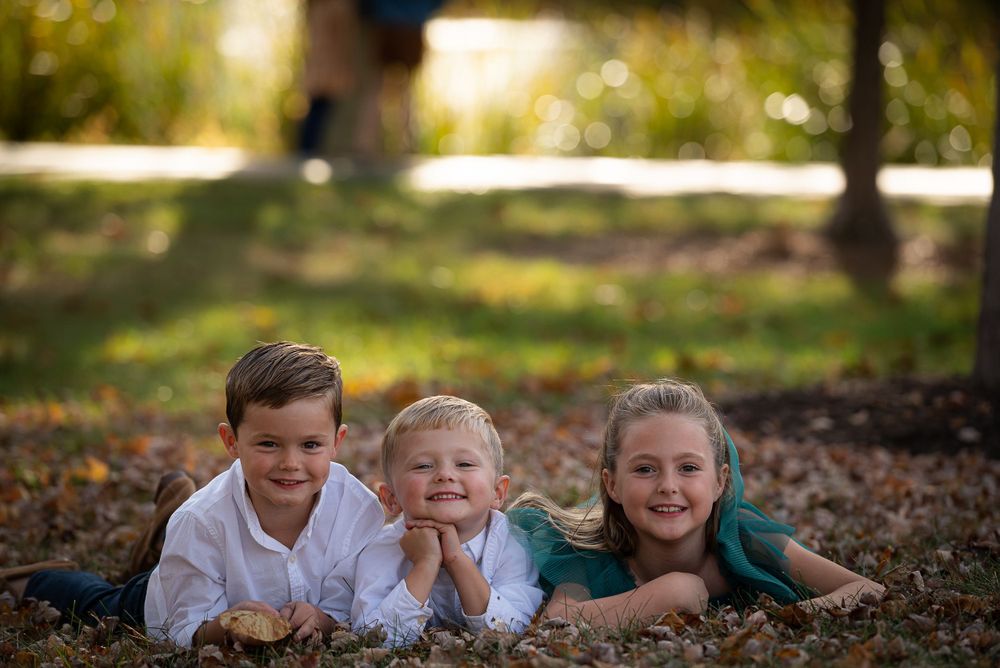Three smiling children lying on autumn leaves in a park.