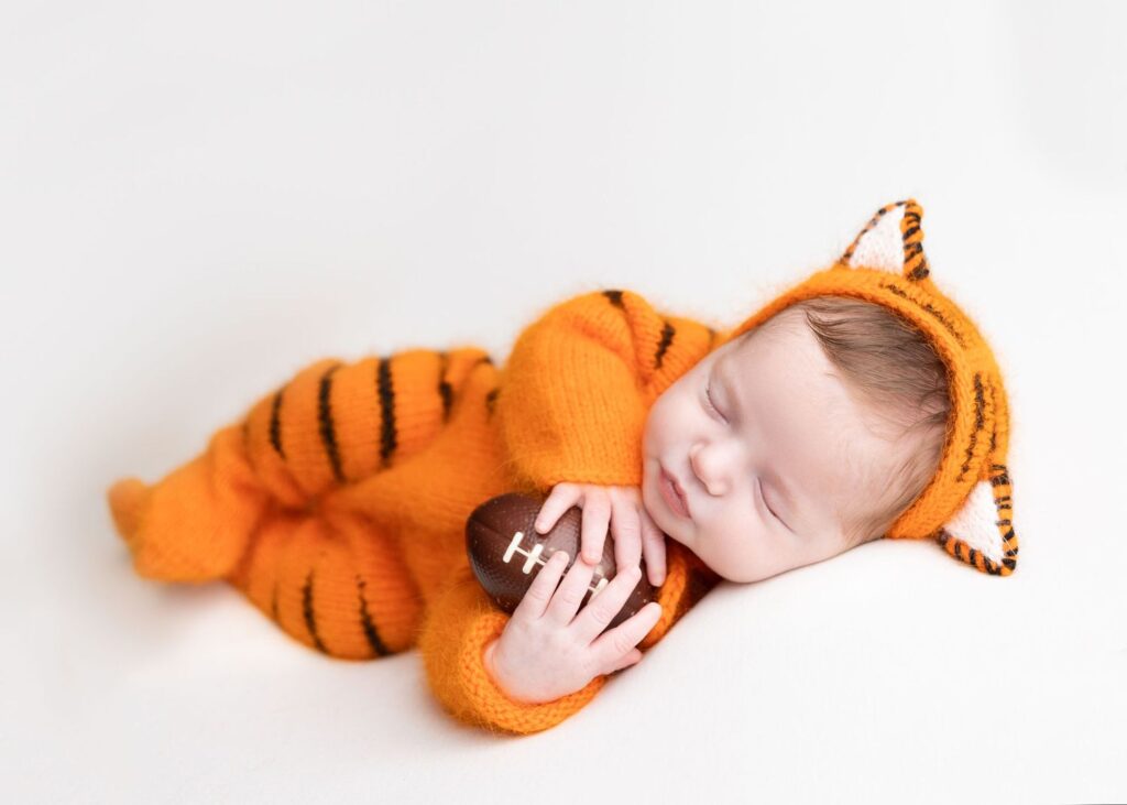 Newborn baby dressed in a tiger costume holding a small football.