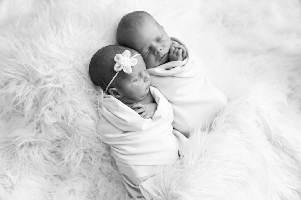 Two newborn babies swaddled and lying close to each other on a fluffy white blanket.