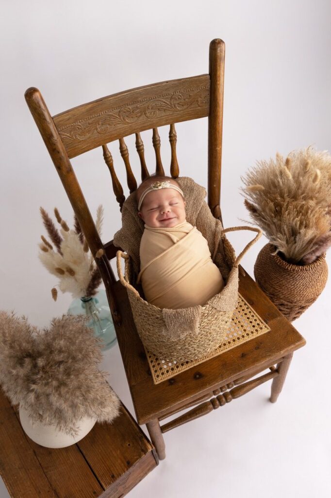 Newborn baby swaddled and smiling while resting on a vintage wooden chair, surrounded by decorative pampas grass.
