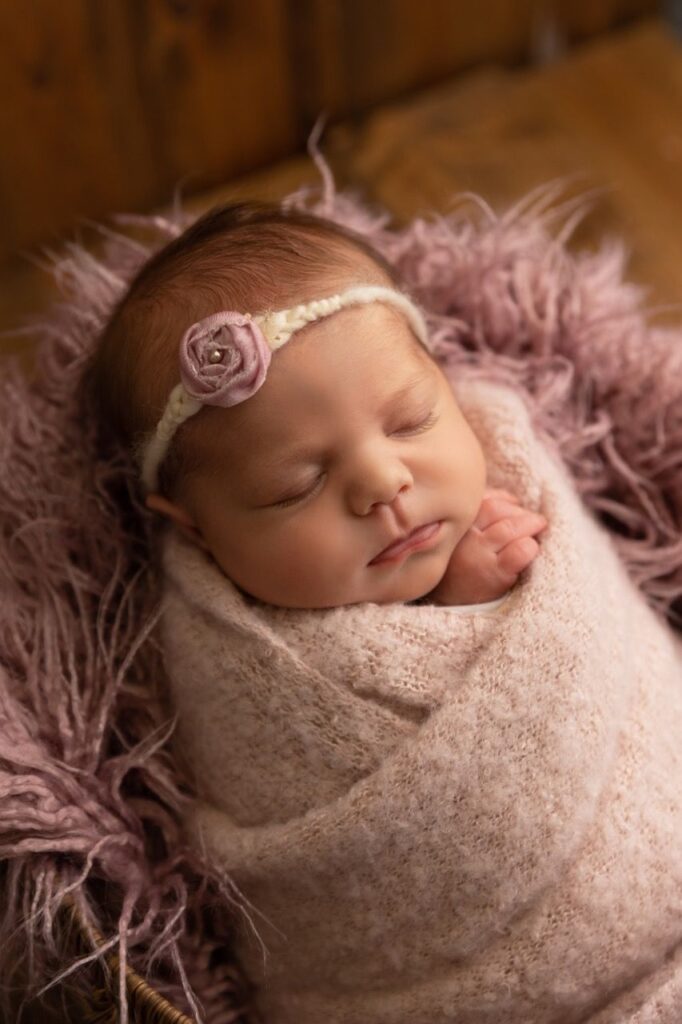 Newborn baby wrapped in a pink blanket, wearing a flower headband, sleeping peacefully.