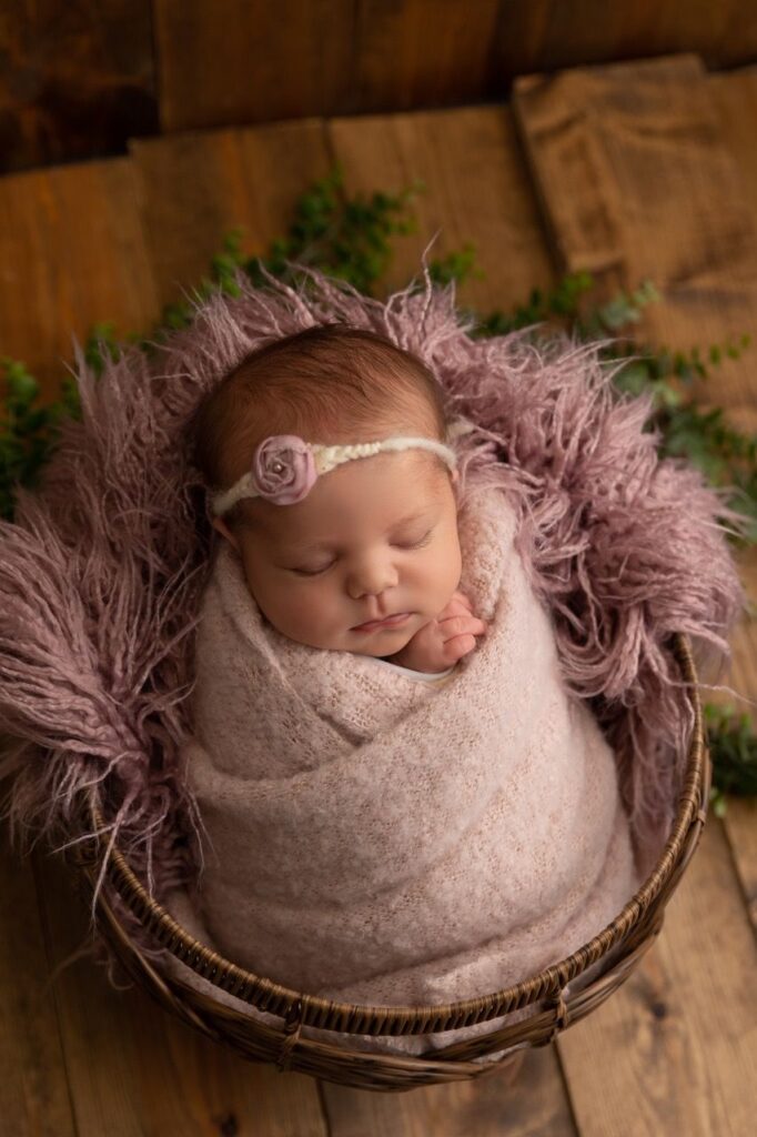 A newborn baby wrapped in a soft pink blanket, resting in a woven basket, with a delicate floral headband.