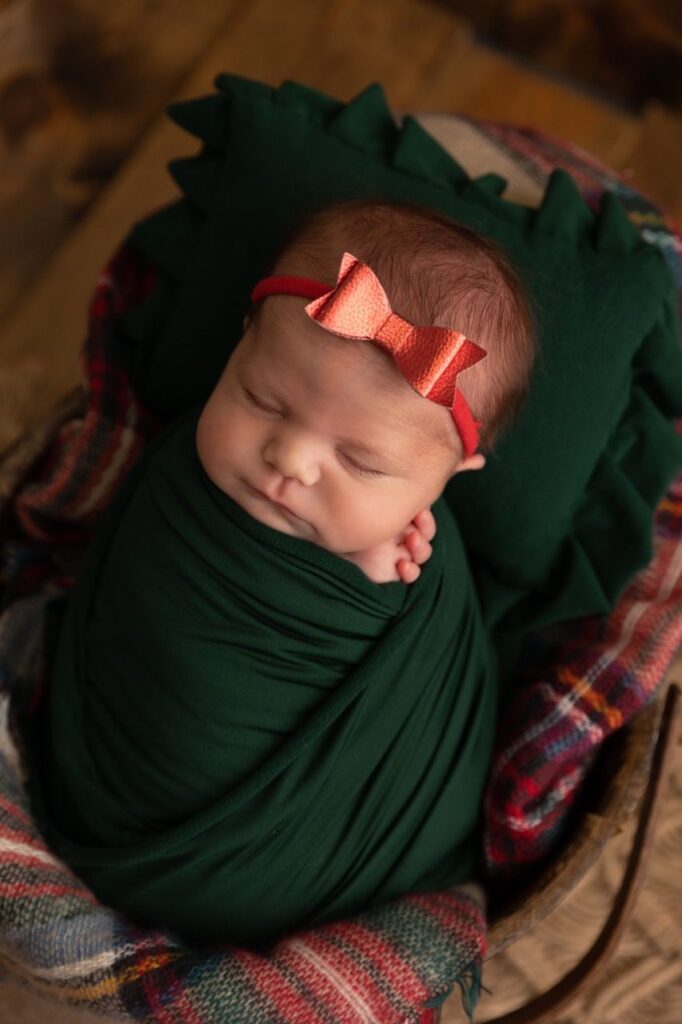 Newborn baby wrapped in green cloth and wearing a red bow headband, resting in a basket.