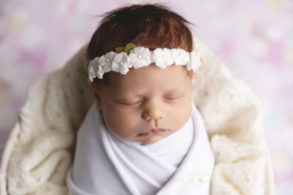 Newborn baby swaddled in a cream blanket and wearing a white floral headband, sleeping peacefully.