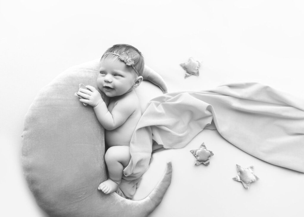 A black and white image of a newborn baby lying on a crescent moon-shaped pillow with a soft blanket and star ornaments.