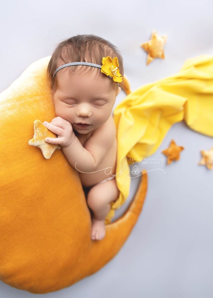 Newborn baby sleeping on a crescent moon prop with star-shaped cushions and a yellow blanket.