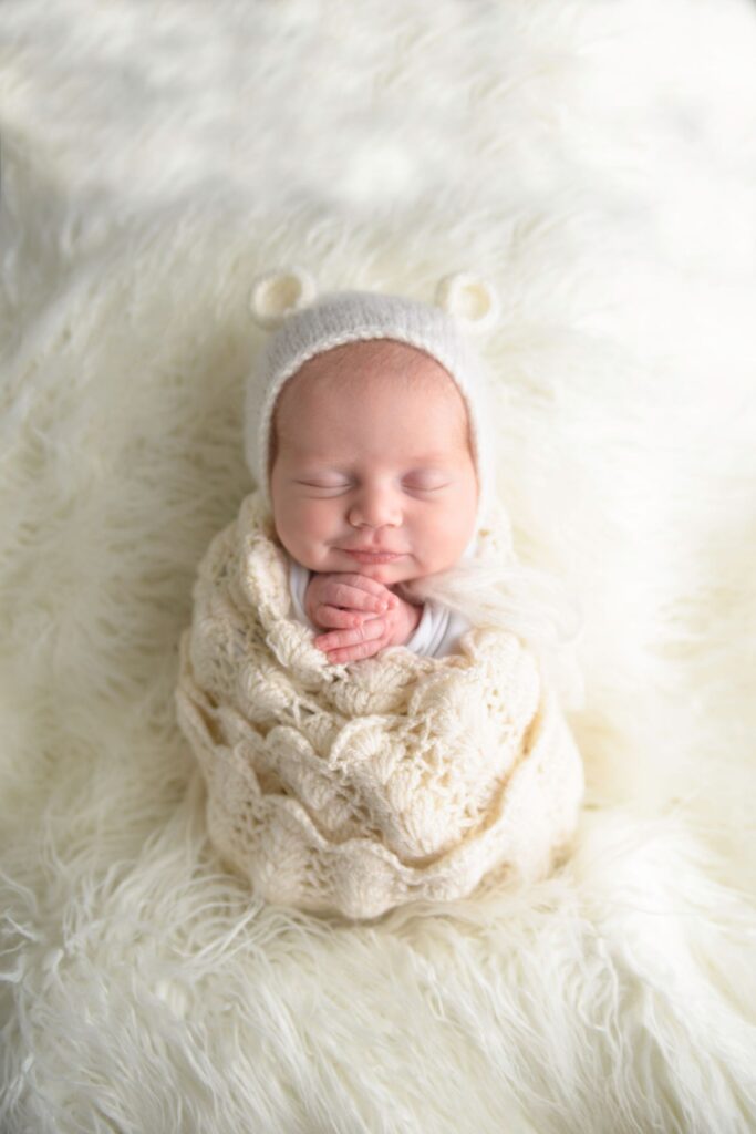 Newborn baby swaddled in a knit blanket on a fluffy white backdrop, wearing a bear-eared beanie.