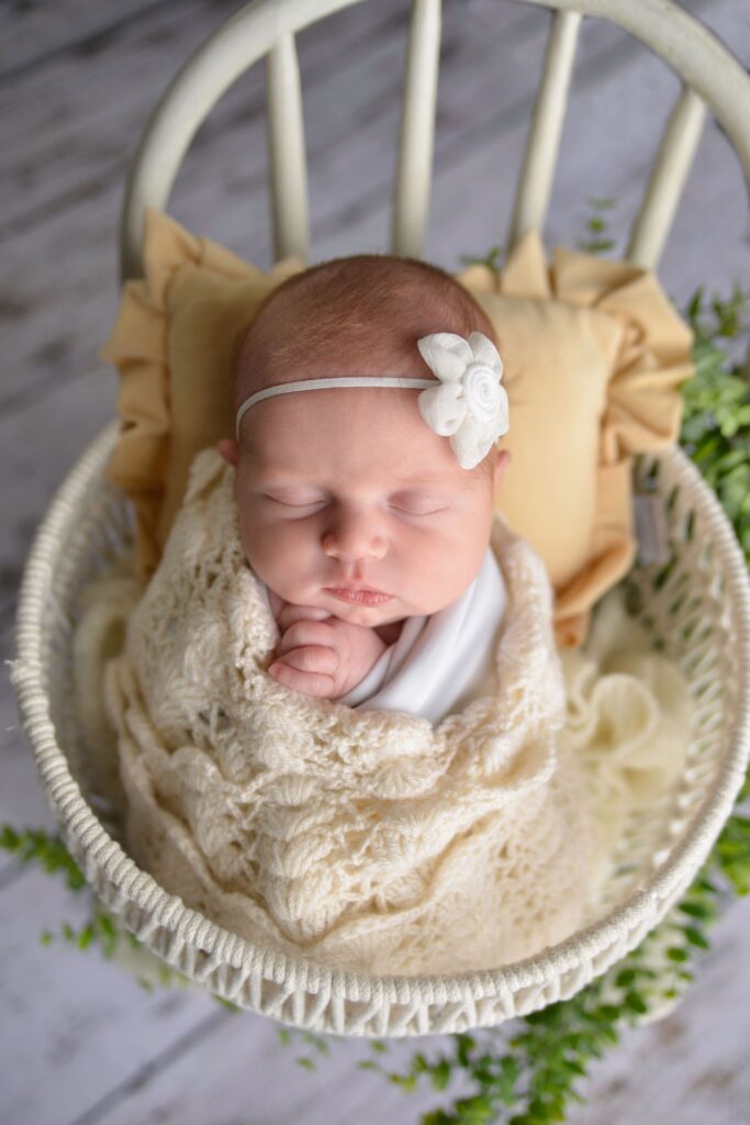 Newborn baby sleeping peacefully in a basket with a cream blanket and a white headband.