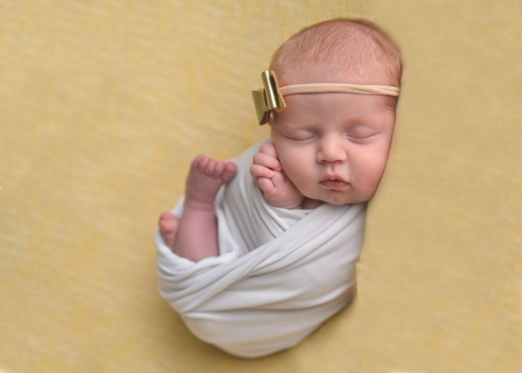 Newborn baby swaddled in white, sleeping peacefully on a yellow blanket with a golden bow headband.