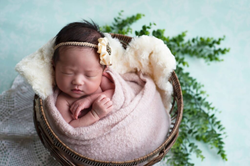 Newborn baby wrapped in a pink blanket and resting in a basket with a floral headband.