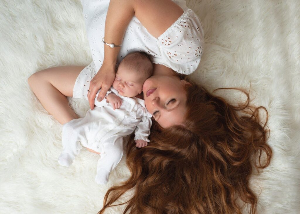 A mother with long, flowing red hair lies on a white furry blanket, cradling her sleeping newborn baby dressed in white. both are in a serene pose, capturing a peaceful, tender moment.
