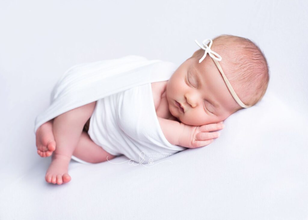 Newborn baby sleeping peacefully with a white bow headband on a soft, light-colored backdrop.