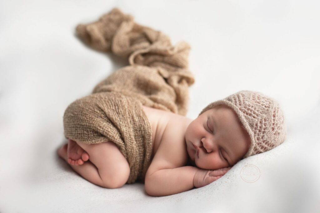 A newborn baby sleeping peacefully wrapped in a beige swaddle with a matching bonnet.