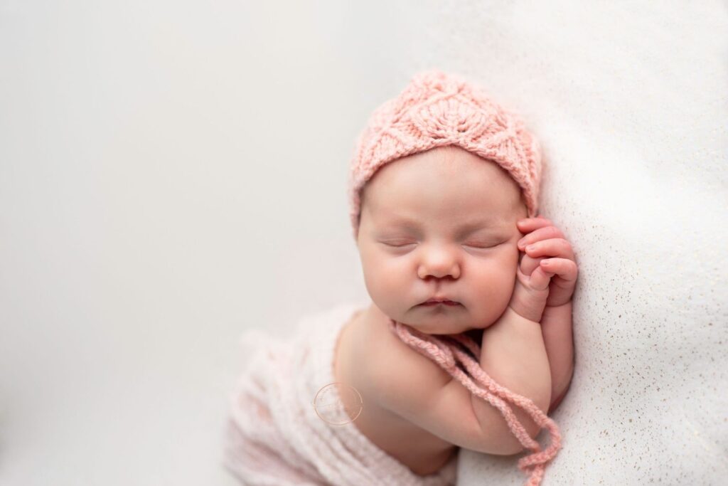 Sleeping newborn in a pink knit hat and wrap against a soft background.