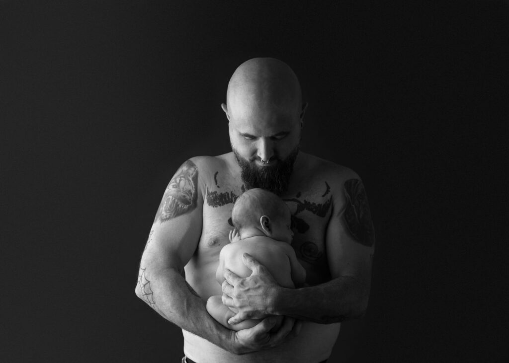 Bearded man with tattoos holding a newborn baby, both in a tender embrace, against a dark background.