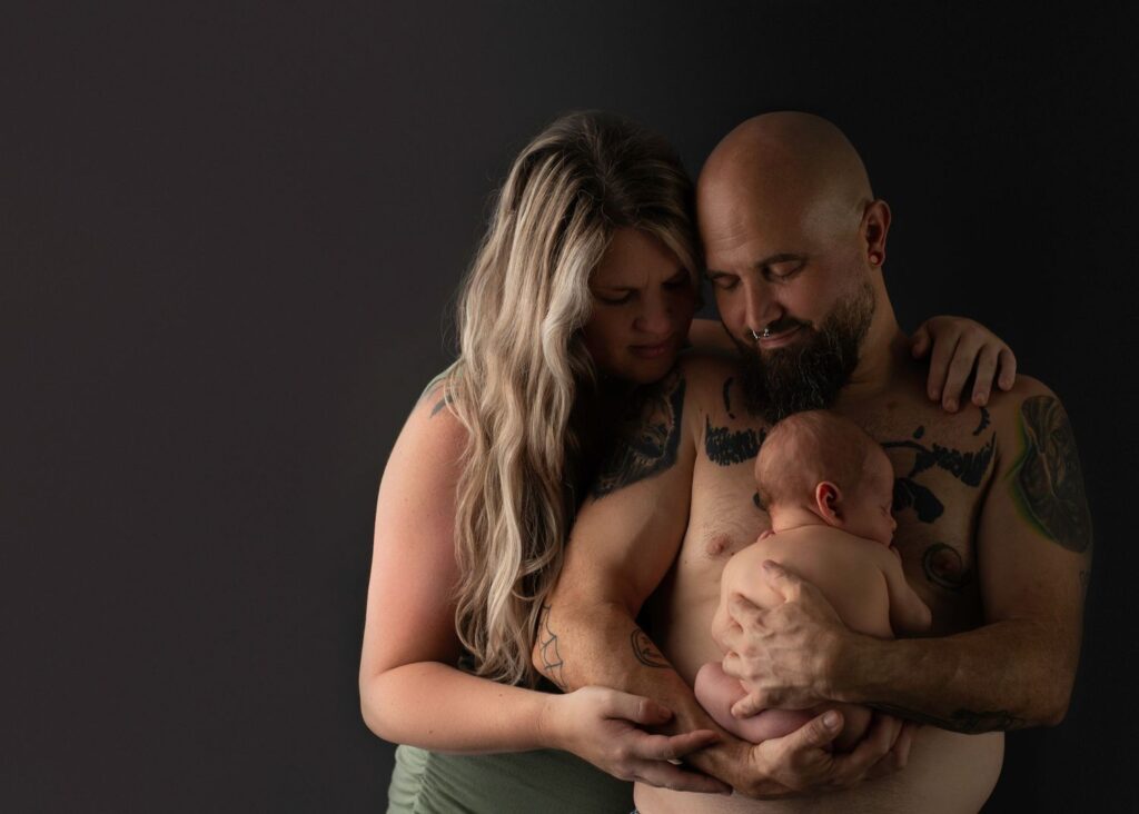 A tender moment as a new dad with tattoos cradles a newborn baby against a dark background, alongside a blonde woman, both showing affection and emotional connection.