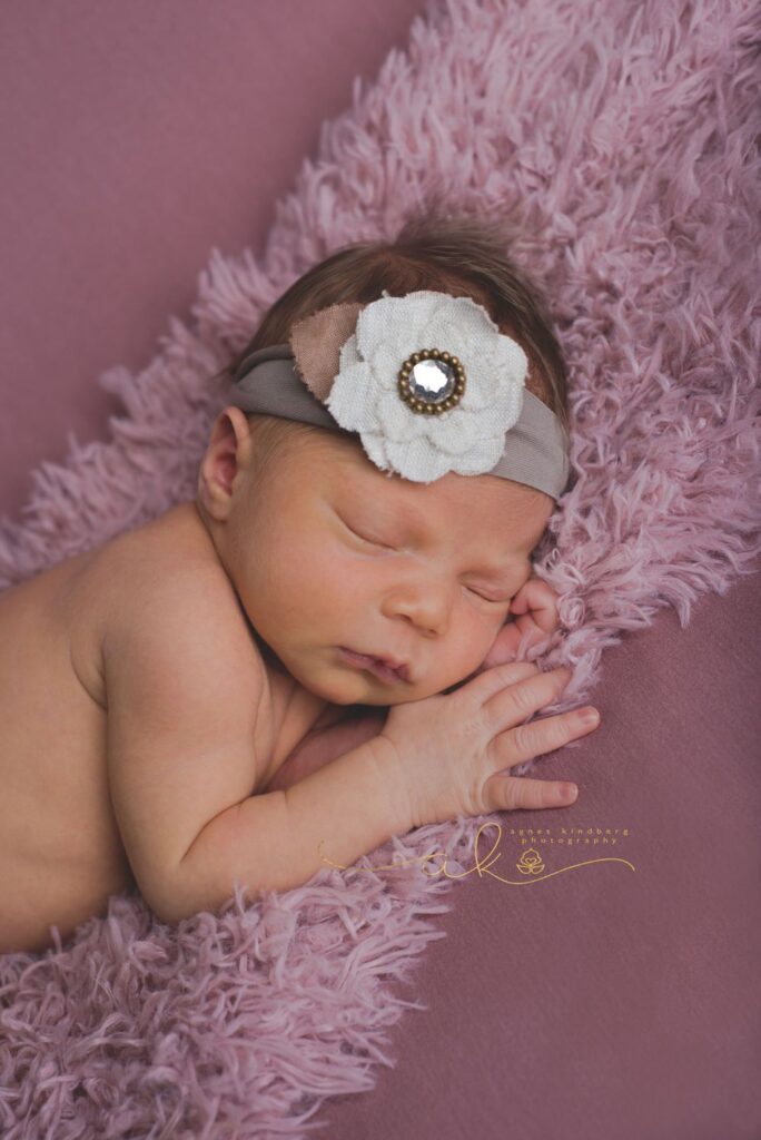 Newborn baby sleeping on a pink fluffy blanket, wearing a headband with a white flower.
