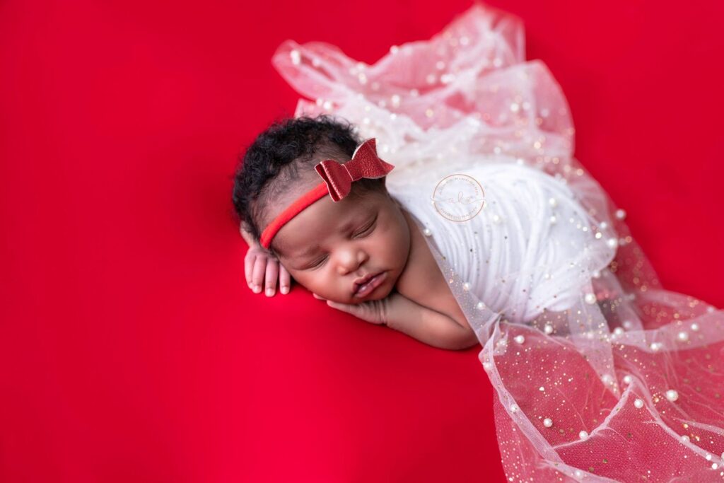 A newborn baby girl sleeping peacefully on a red background, dressed in a white outfit with a delicate fabric and wearing a red bow on her head.