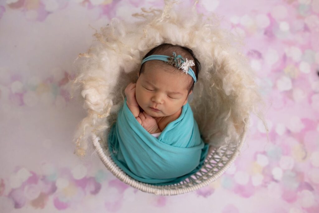 Newborn baby swaddled in blue, resting in a white fluffy basket with a floral backdrop.