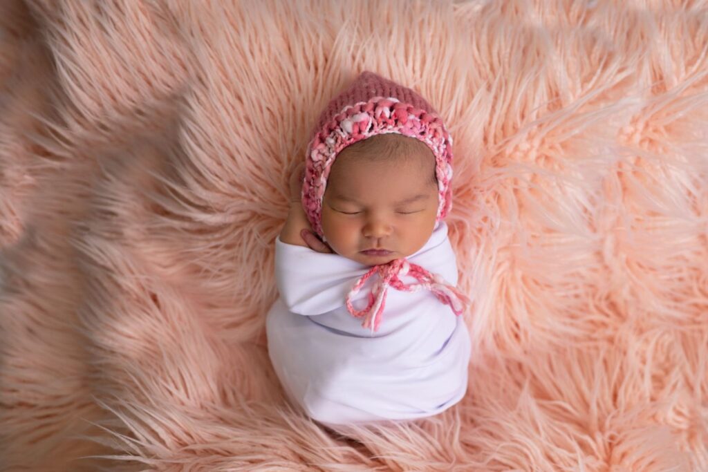 A newborn baby swaddled in white, wearing a pink knit hat, sleeping on a fluffy pink blanket.