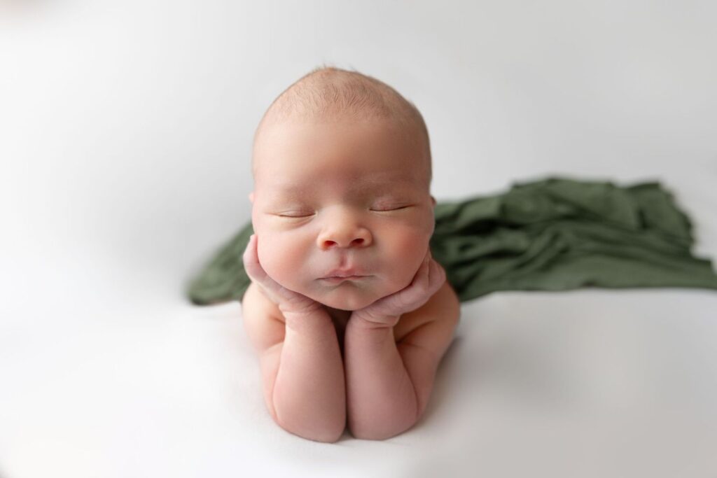 Newborn baby resting its head on its hands while asleep on a white surface with a green cloth draped behind.