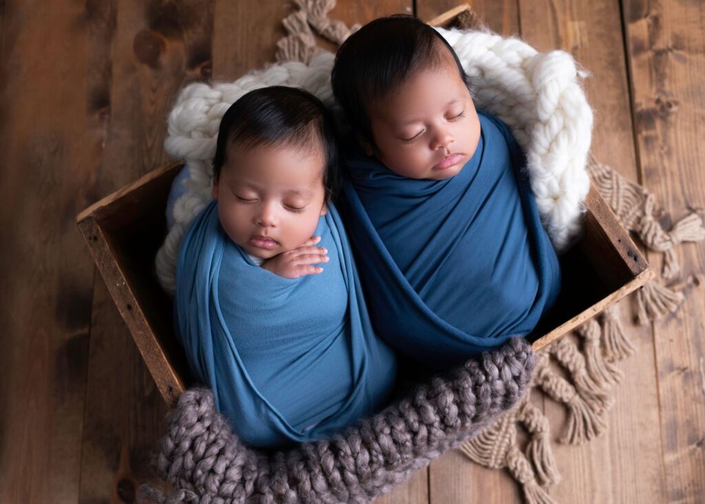 Twin infants swaddled in blue, sleeping peacefully in wooden crates with soft knitted blankets.