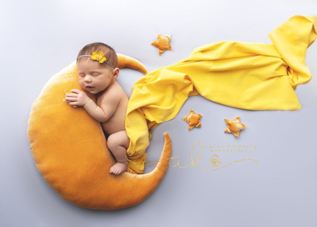 Newborn baby asleep on a crescent moon pillow with yellow stars and fabric accentuating the dreamy setup.